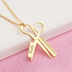 May include: Gold-colored necklace with a pendant featuring scissors and a comb, a design often associated with hairstylists. The pendant has the name "Ruth" engraved on it. The necklace is displayed on a white and pink surface.