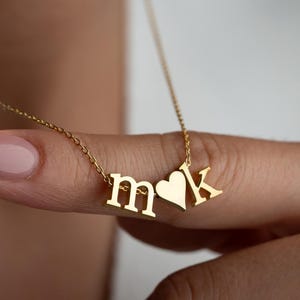 May include: A gold-colored necklace with the letters "m" and "k" and a heart-shaped charm. The necklace is on a delicate chain. The letters and heart are in a sans-serif font. The necklace is being held up by a person's finger.