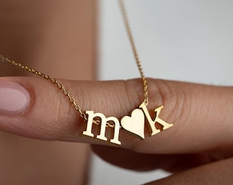 May include: A gold-colored necklace with the letters "m" and "k" and a heart-shaped charm. The necklace is on a delicate chain. The letters and heart are in a sans-serif font. The necklace is being held up by a person's finger.