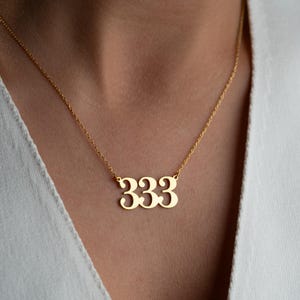 Puede incluir: Un collar dorado con un colgante con el n&uacute;mero 333. El colgante est&aacute; suspendido de una fina cadena. El collar se lleva sobre una parte superior blanca.