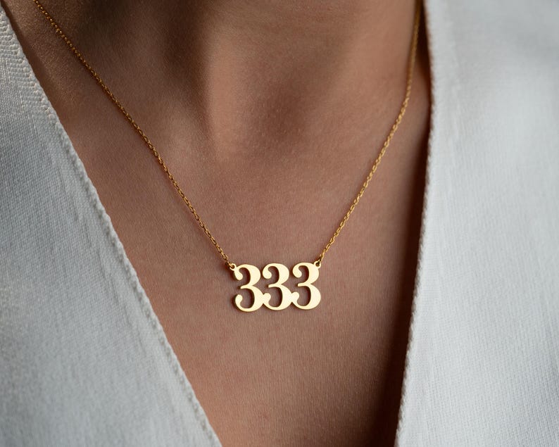 Puede incluir: Un collar dorado con un colgante con el n&uacute;mero 333. El colgante est&aacute; suspendido de una fina cadena. El collar se lleva sobre una parte superior blanca.