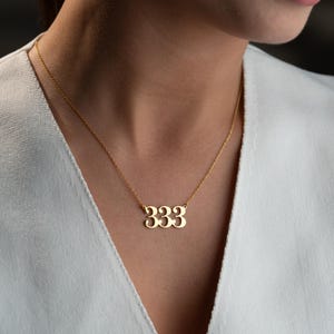 Puede incluir: Un collar dorado con un colgante con el n&uacute;mero "333". El collar lo lleva una persona con una parte superior blanca. El colgante est&aacute; hecho de un metal brillante y cuelga de una cadena delicada.