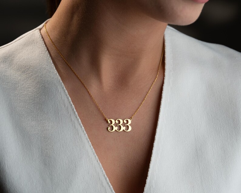 Puede incluir: Un collar dorado con un colgante con el n&uacute;mero "333". El collar lo lleva una persona con una parte superior blanca. El colgante est&aacute; hecho de un metal brillante y cuelga de una cadena delicada.