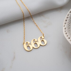 Puede incluir: Un collar dorado con el n&uacute;mero "666" como colgante. El collar est&aacute; en una cadena dorada y se muestra sobre una superficie blanca. Es una pieza de joyer&iacute;a.