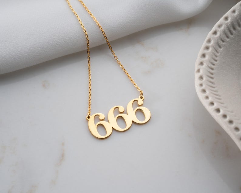 Puede incluir: Un collar dorado con el n&uacute;mero "666" como colgante. El collar est&aacute; en una cadena dorada y se muestra sobre una superficie blanca. Es una pieza de joyer&iacute;a.