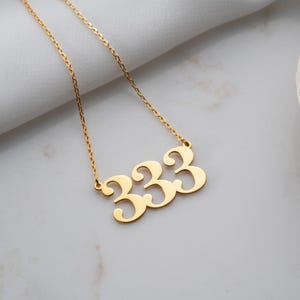 Puede incluir: Un collar dorado con un colgante con el n&uacute;mero 333. El colgante, con una fuente estilizada, cuelga de una fina cadena. El collar se muestra sobre una superficie blanca.
