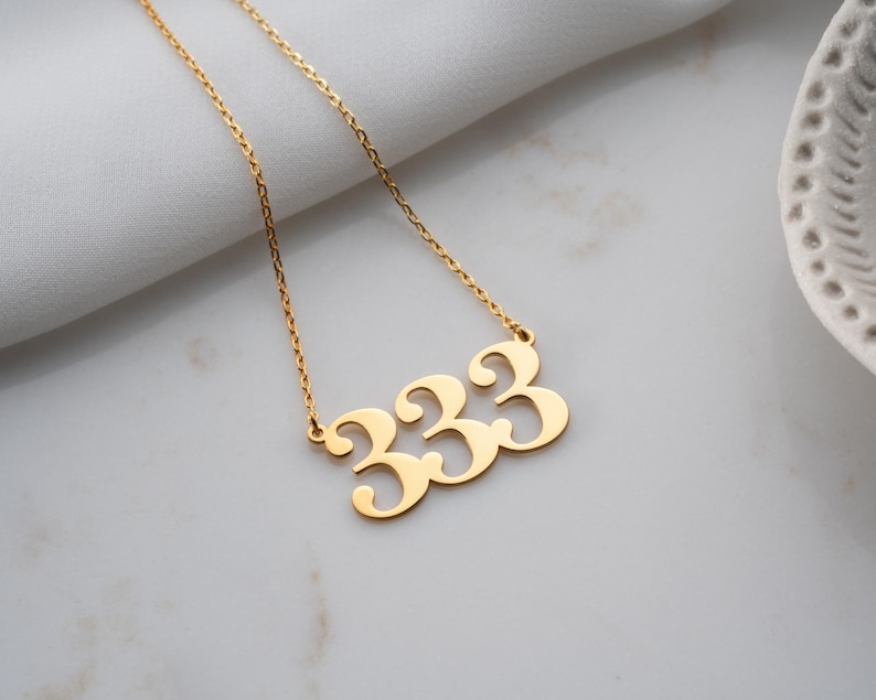 Puede incluir: Un collar dorado con un colgante con el n&uacute;mero 333. El colgante, con una fuente estilizada, cuelga de una fina cadena. El collar se muestra sobre una superficie blanca.