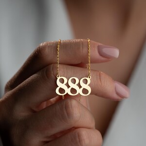 Puede incluir: Un collar dorado con un colgante con el n&uacute;mero 888. El colgante cuelga de una fina cadena. El collar lo sostiene una persona con las u&ntilde;as cuidadas, lo que sugiere un inter&eacute;s por la joyer&iacute;a y la moda.