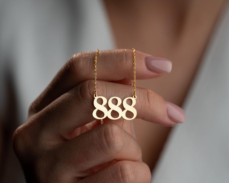 Puede incluir: Un collar dorado con un colgante con el n&uacute;mero 888. El colgante cuelga de una fina cadena. El collar lo sostiene una persona con las u&ntilde;as cuidadas, lo que sugiere un inter&eacute;s por la joyer&iacute;a y la moda.