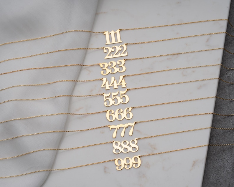 Puede incluir: Una colecci&oacute;n de collares dorados con n&uacute;meros, cada uno con un n&uacute;mero diferente de tres d&iacute;gitos del 111 al 999. Los collares se exhiben sobre una superficie de m&aacute;rmol blanco y gris, mostrando las delicadas cadenas y dise&ntilde;os de colgantes.