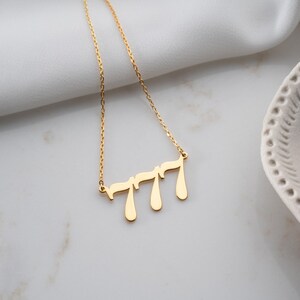 Puede incluir: Un collar dorado con una cadena delicada. El colgante presenta el n&uacute;mero "777" en una fuente estilizada y fluida. El collar se muestra sobre una superficie blanca, con un fondo borroso.