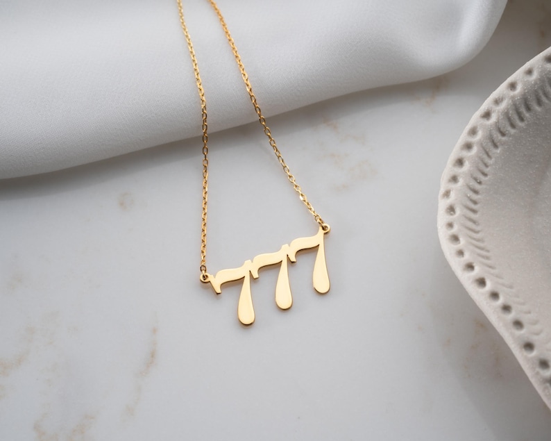 Puede incluir: Un collar dorado con una cadena delicada. El colgante presenta el n&uacute;mero "777" en una fuente estilizada y fluida. El collar se muestra sobre una superficie blanca, con un fondo borroso.