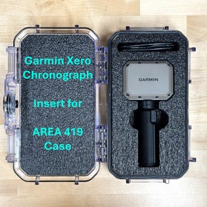 May include: A clear plastic case with a black foam insert for a Garmin Xero Chronograph. The case is open and the insert is empty. The text "Garmin Xero Chronograph Insert for AREA 419 Case" is printed on the foam.