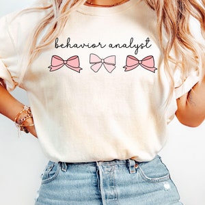 Pode incluir: Uma camiseta branca com o texto "behavior analyst" impresso nela. Três laços rosa estão centralizados abaixo do texto.