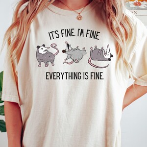Puede incluir: Una camiseta blanca con un gr&aacute;fico de tres zarig&uuml;eyas en diferentes poses. El texto "It's fine. I'm fine. Everything is fine." est&aacute; impreso en la camiseta.