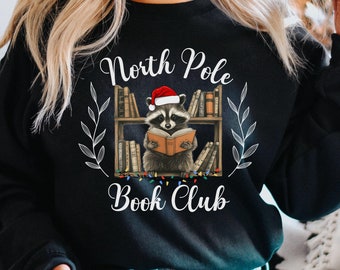 May include: Black sweatshirt with a graphic of a raccoon wearing a Santa hat and reading a book on a bookshelf. The text "North Pole Book Club" is printed on the sweatshirt.