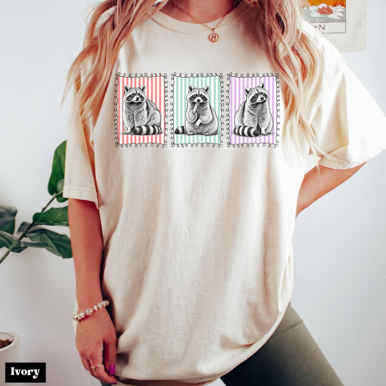 Op de afbeelding: Ivoor kleurig T-shirt met een afbeelding van drie wasberen in kaders met kleurrijke gestreepte achtergronden. De wasberen hebben zwart-wit gestreepte staarten. Het shirt heeft een relaxte pasvorm. Het woord "Ivory" staat onderaan.