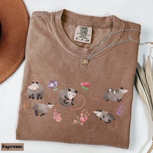 May include: Espresso-colored t-shirt with a crew neck, featuring a whimsical design of opossums and flowers. The Comfort Colors brand label is visible. A silver necklace with a pendant is draped across the shirt.