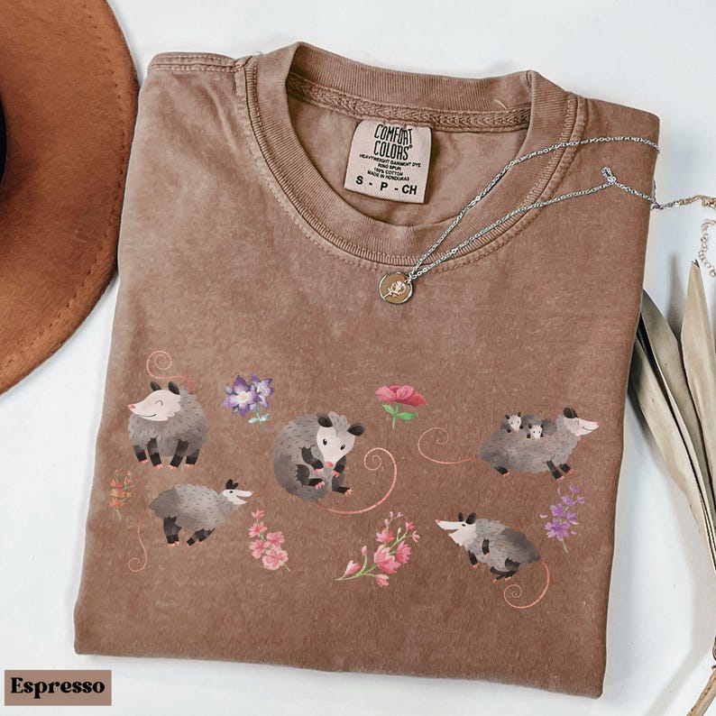 May include: Espresso-colored t-shirt with a crew neck, featuring a whimsical design of opossums and flowers. The Comfort Colors brand label is visible. A silver necklace with a pendant is draped across the shirt.