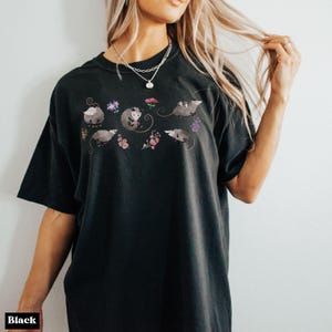 May include: Black t-shirt featuring a whimsical design of opossums and flowers. The opossums are depicted in various poses, surrounded by delicate floral elements in shades of pink, purple, and white. The shirt is a comfortable, casual style.