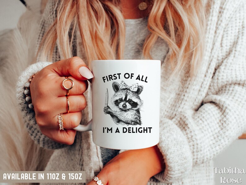 May include: White ceramic mug with a black and white illustration of a raccoon wearing a bow and holding a knife. The text "First of all I'm a delight" is printed on the mug.