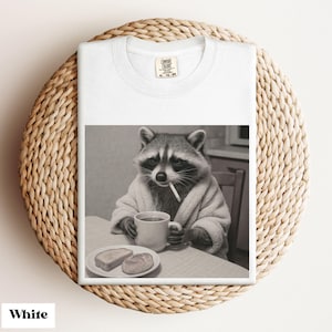 May include: White t-shirt featuring a black and white image of a raccoon in a bathrobe, smoking a cigarette, and drinking from a mug. A plate with toast is on the table. The shirt has a crew neck and a tag that reads "COMFORT COLORS".