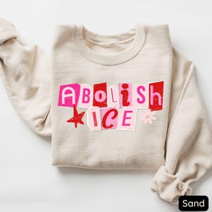 May include: Sand-colored sweatshirt with the words "Abolish ICE" in a collage-style design using pink and red letters. A red star and a small flower accent the text. The sweatshirt is folded neatly.