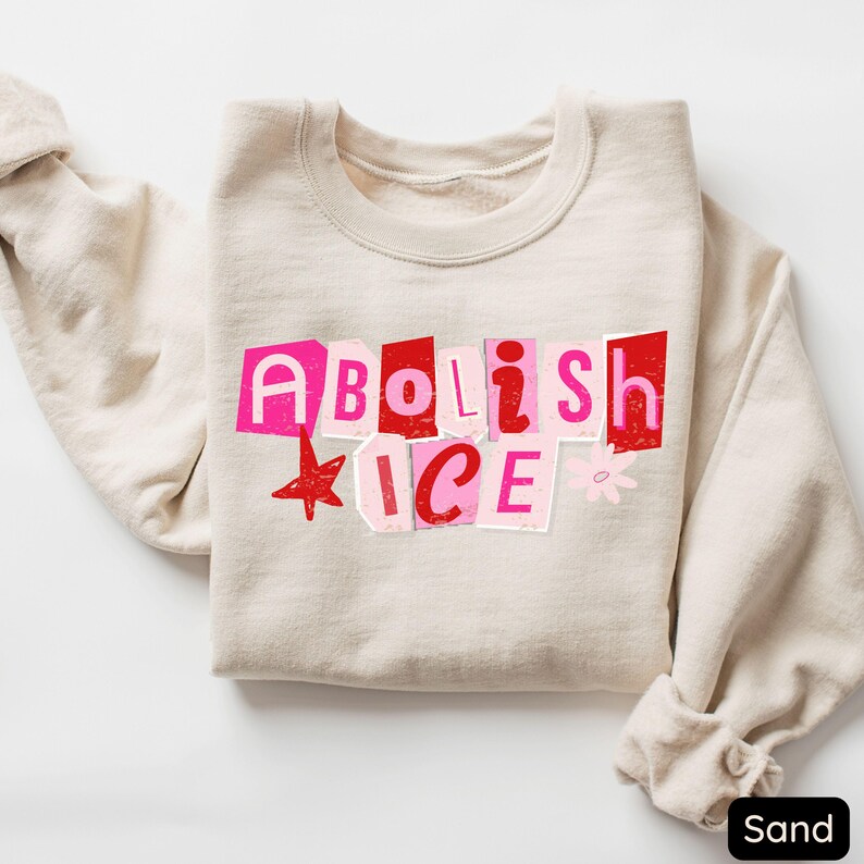 May include: Sand-colored sweatshirt with the words "Abolish ICE" in a collage-style design using pink and red letters. A red star and a small flower accent the text. The sweatshirt is folded neatly.