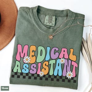 Op de afbeelding: Een groen T-shirt met de tekst "MEDICAL ASSISTANT" in kleurrijke, retro-stijl letters met madeliefjes. De letters zijn zwart omrand en hebben een geruit patroon eronder.