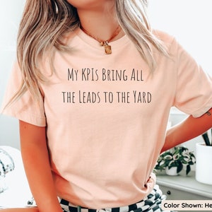 Pu&ograve; includere: Una t-shirt color pesca con il testo "My KPIs bring all the leads to the yard" stampato sul davanti. La t-shirt &egrave; mostrata su una modella che indossa una gonna a quadri bianchi e neri. Colore mostrato: Heather Peach.