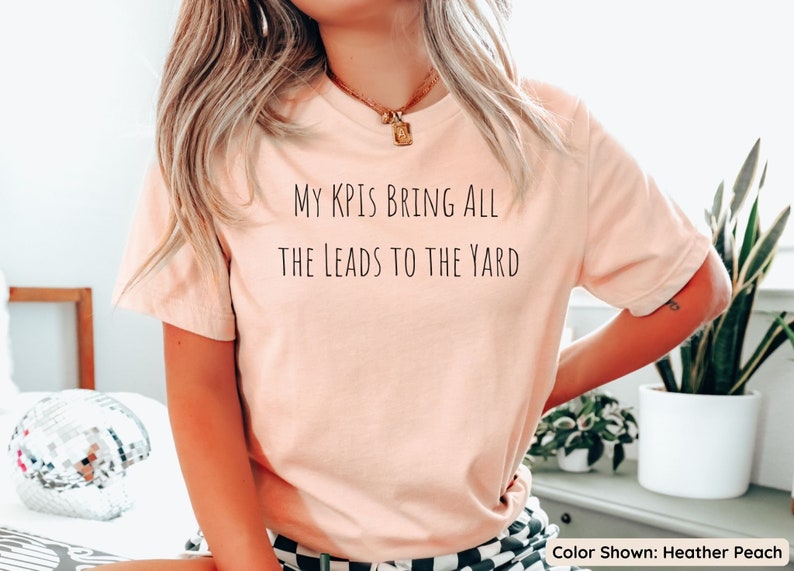 Pu&ograve; includere: Una t-shirt color pesca con il testo "My KPIs bring all the leads to the yard" stampato sul davanti. La t-shirt &egrave; mostrata su una modella che indossa una gonna a quadri bianchi e neri. Colore mostrato: Heather Peach.