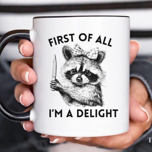 May include: White ceramic mug with a black handle. The mug has a black and white illustration of a raccoon wearing a bow and holding a knife. The text "First of all I'm a delight" is printed on the mug.