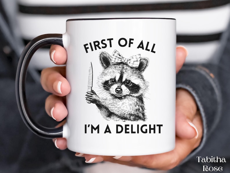 May include: White ceramic mug with a black handle. The mug has a black and white illustration of a raccoon wearing a bow and holding a knife. The text "First of all I'm a delight" is printed on the mug.