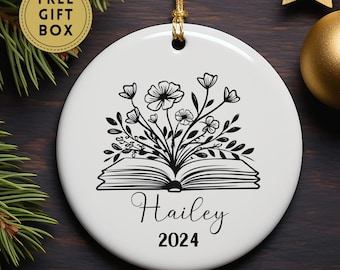May include: White ceramic ornament with a black and white illustration of a book with flowers growing out of it. The text "Hailey 2024" is printed below the book.