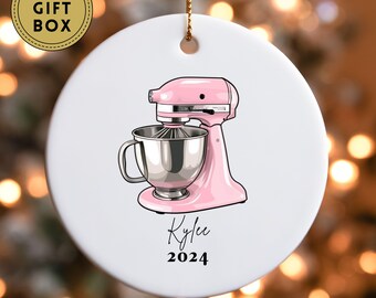 May include: A white ceramic Christmas ornament with a pink stand mixer and the text "Kylee 2024".