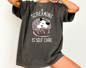 May include: A dark gray t-shirt with a graphic of a cartoon possum with the text "Screaming is Self Care".
