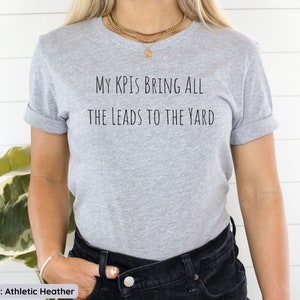 Pu&ograve; includere: Una maglietta grigia con la scritta "My KPIs bring all the leads to the yard" stampata sul davanti.
