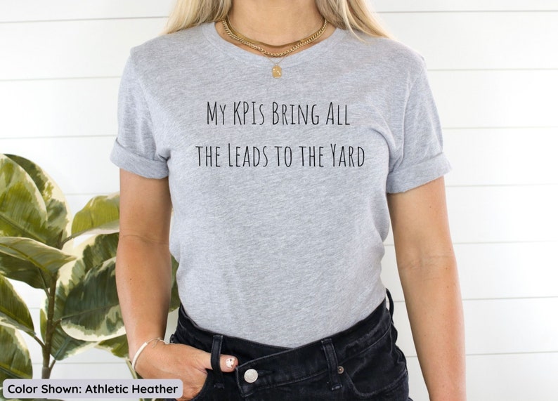 Pu&ograve; includere: Una maglietta grigia con la scritta "My KPIs bring all the leads to the yard" stampata sul davanti.