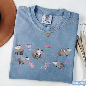 May include: Blue jean colored Comfort Colors t-shirt featuring a graphic of several opossums and flowers. The shirt has a crew neck and short sleeves. A gold necklace with a pendant is draped across the shirt.