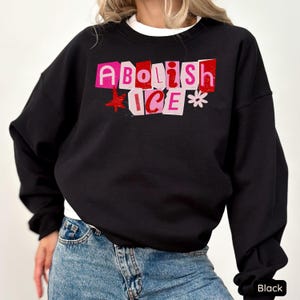 May include: Black sweatshirt with the text "Abolish ICE" in pink and red block letters. A red star and a white flower accent the text. The sweatshirt is a crew neck style.