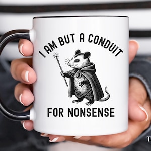 Puede incluir: Taza de cer&aacute;mica blanca con asa negra. La taza presenta una ilustraci&oacute;n en blanco y negro de una zarig&uuml;eya sosteniendo una varita y el texto "Solo soy un conducto para tonter&iacute;as".