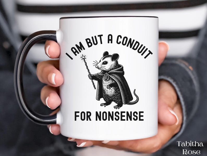 Puede incluir: Taza de cer&aacute;mica blanca con asa negra. La taza presenta una ilustraci&oacute;n en blanco y negro de una zarig&uuml;eya sosteniendo una varita y el texto "Solo soy un conducto para tonter&iacute;as".