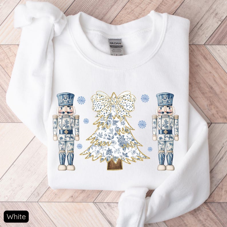 May include: White sweatshirt with a blue and gold Christmas tree and two nutcracker soldiers on either side. The tree is decorated with gold glitter and blue and white floral patterns. The nutcrackers are wearing blue and white uniforms with gold accents.