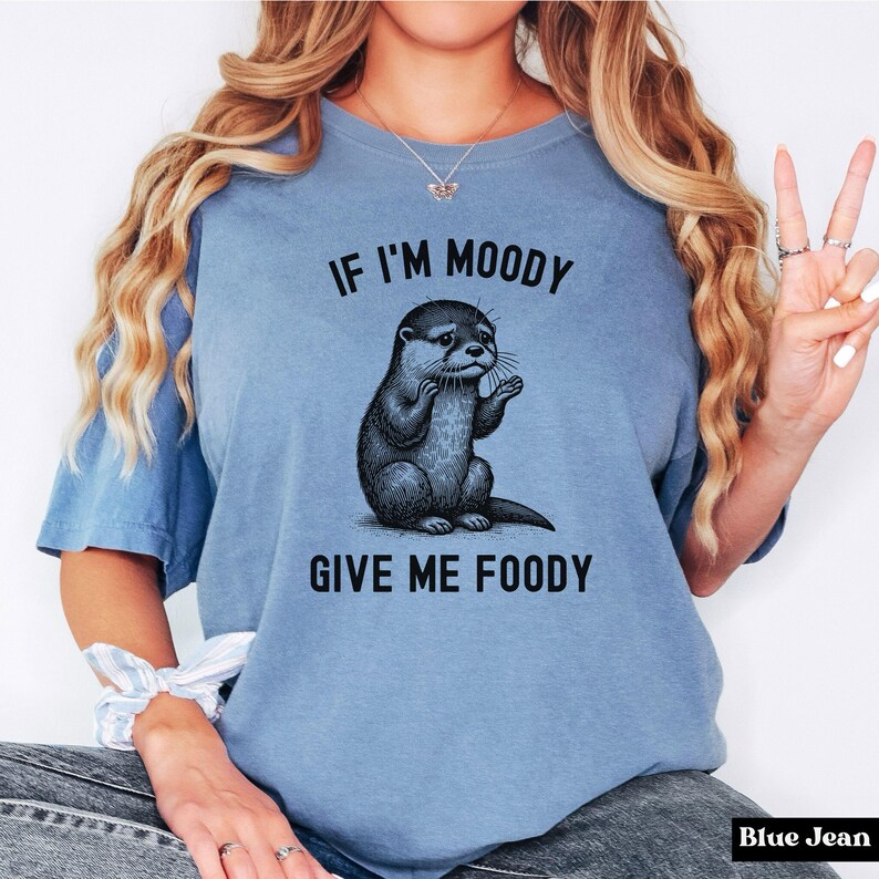 May include: A blue t-shirt with a graphic of an otter and the text "If I'm Moody Give Me Foody".