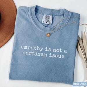Peut inclure: T-shirt bleu jean avec l'inscription blanche "empathy is not a partisan issue". Un collier doré avec un pendentif est posé sur le t-shirt. Le t-shirt porte une étiquette Comfort Colors.