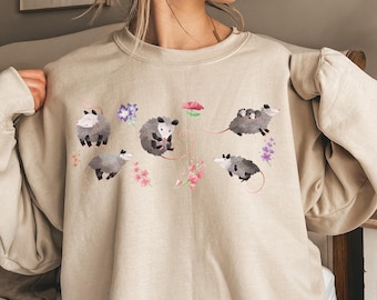 May include: A sand-coloured sweatshirt with a playful design of opossums and floral elements. The opossums are illustrated in different poses, set amongst watercolour-style flowers in pink, purple, and blue. The word "Sand" is printed in a black rectangle.