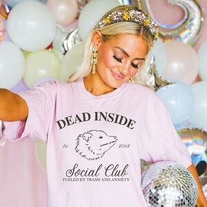 May include: Pink t-shirt with the text "Dead Inside Social Club Fueled by Trash and Anxiety" and a black and white illustration of an opossum. The text "Est. 2024" is also on the shirt.