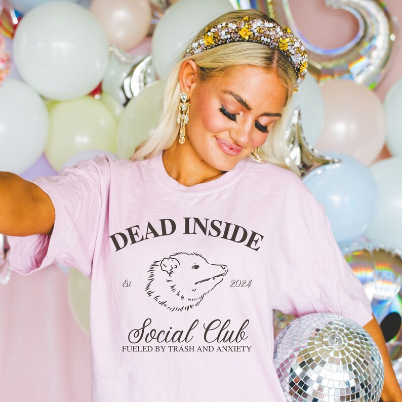 May include: Pink t-shirt with the text "Dead Inside Social Club Fueled by Trash and Anxiety" and a black and white illustration of an opossum. The text "Est. 2024" is also on the shirt.