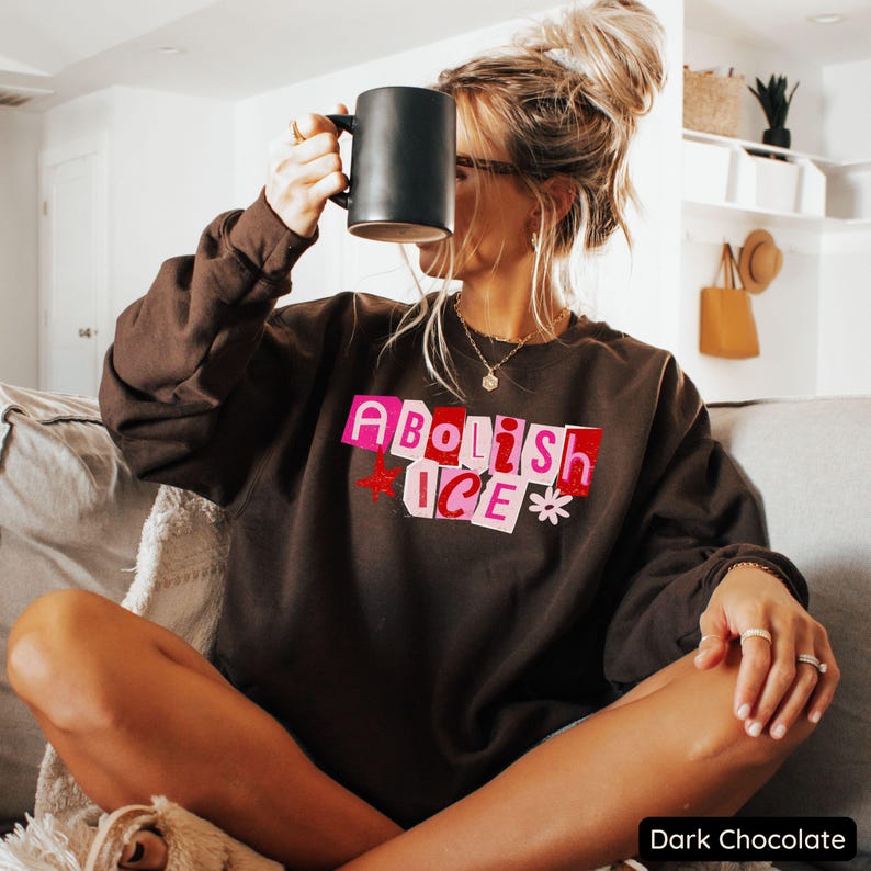 May include: Dark chocolate-colored sweatshirt with the text "Abolish ICE" in pink and red block letters. The sweatshirt features a crew neck and long sleeves. A black mug is held up to the face. The bottom right corner of the image has the text "Dark Chocolate".