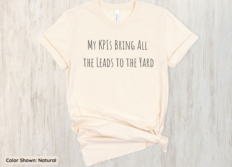 Pu&ograve; includere: Una t-shirt di colore naturale con la scritta "My KPIs bring all the leads to the yard" stampata sul davanti.